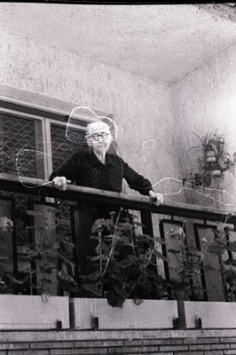 A monochrome image captures an older woman on a balcony. She wears a dark, buttoned shirt and stands with hands on the railin...