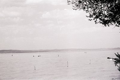 A wide coastal view shows figures wading and boating in calm water, with a low, distant shoreline visible through a hazy atmo...