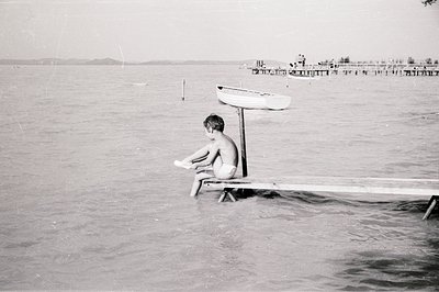 A young boy sits pensively on a pier, partially submerged in water. A small boat is secured overhead. Visible in the distance...