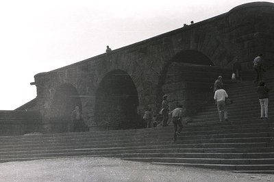 Striking black & white image of tiered stone steps ascending to a monumental, arched structure, possibly a fortress or public...