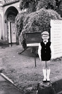A young boy, likely in the 1960s, stands proudly atop a low stone pedestal. He wears a formal outfit: a bow tie, vest, collar...