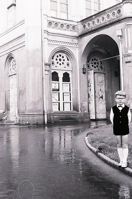 A young boy in a dark vest and knee socks stands on a wet road, with an ornate building facade visible behind him. Architectu...