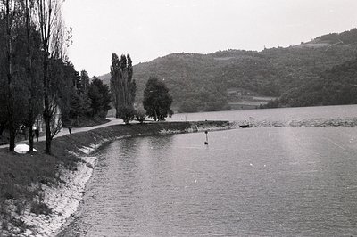 A view of a still lake framed by a grassy bank and distant, forested hills. A paved path borders the water, with a few figure...