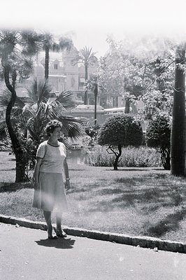 A woman in a simple dress and sandals stands beside a paved road, framed by lush tropical foliage and a waterway. A grand, or...