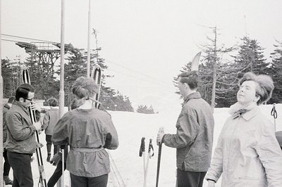 Four figures stand amidst snow-covered evergreens, near a ski lift tower. Two men, one smoking, and two women in 1960s winter...