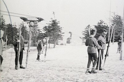 Vintage photo depicts skiers at a rope tow lift, likely in the 1940s or 50s. Several individuals are poised to ride or disemb...