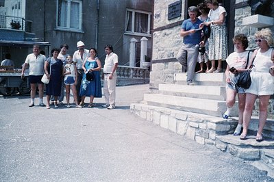 A group gathers on steps outside a stone building; architecture suggests a European coastal setting. Fashion trends indicate ...