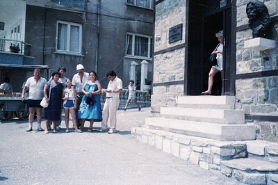 A group of tourists, dressed in 1970s attire, poses outside a stone building. A bust of a man sits atop the building's entry....