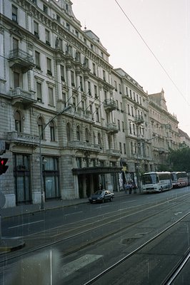 A wide, multistory building with ornate balconies lines a city street. Tram tracks run along the asphalt road. A bus and car ...