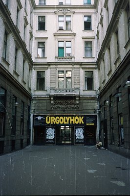 A long, narrow courtyard leads to a storefront entrance in a densely built urban area. The architecture features classical de...