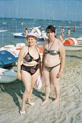 Two women stand on a sandy beach, both in floral-patterned swimsuits. Background shows a crowded shoreline with boats and peo...