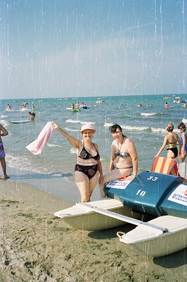 Two women in vintage swimwear pose on a lifeguard beach patrol vehicle. Sandy beach & turquoise water stretch into the backgr...