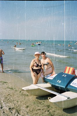 Two women in vintage swimsuits stand beside a small, numbered hydrofoil boat on a sandy beach. The scene includes a wide expa...