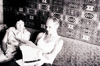 A man engrossed in reading a newspaper, seated on a patterned sofa. A woman is partially visible, seated behind him, appearin...