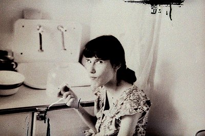 A young woman in a patterned blouse stands in a kitchen, pouring a liquid from a white pitcher. Mid-century modern kitchen fe...