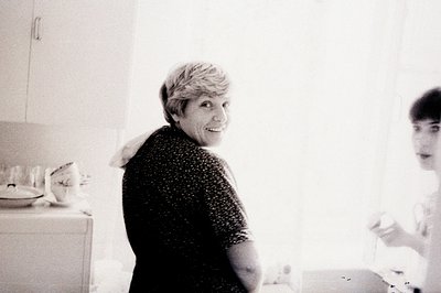 A smiling woman in a floral-patterned top looks back over her shoulder, while a younger person stands in profile near a kitch...