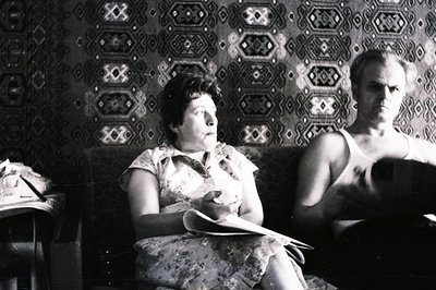 A woman seated on a sofa, holding a document, is framed by a vibrant, patterned wallpaper. A man sits beside her, appearing c...