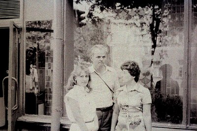 A grayscale, likely candid portrait, captures a man flanked by two young girls in front of a mirrored storefront. The man, li...