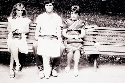 Three young people sit on a weathered park bench. The man, centered, holds a sheet of paper; the two women are on either side...