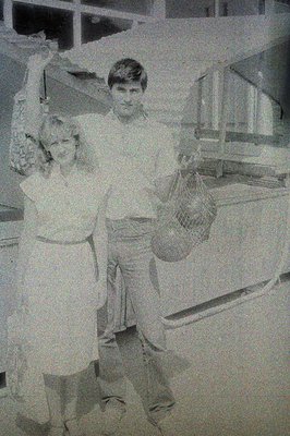 Young man and woman, likely fishermen, stand near a seafood display in what appears to be a market. She wears a short dress; ...
