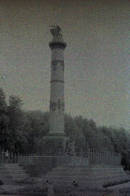 Monumental column with visible relief carvings rises from a stone base and ironwork fence, set against a backdrop of dense fo...