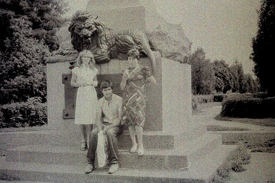Three young people pose near a large lion statue within a tiered, formal garden setting. The statue, flanked by plaques, sits...
