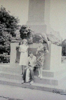 A black and white photo shows three people posed beside an ornate stone lion statue. Two teens sit on the steps, one girl sta...