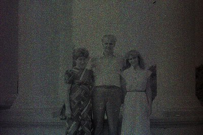Three people stand posing outdoors, likely in front of a building. The woman on the left wears a patterned dress, the man is ...