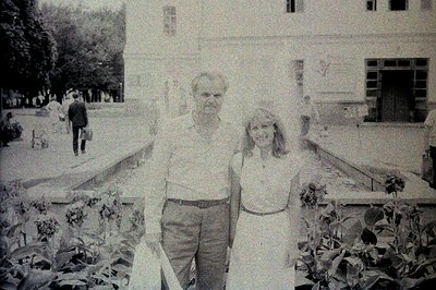 A black & white snapshot depicts a couple posing formally in front of a large, classically styled building with a prominent p...