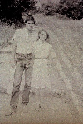 A young man and girl stand outdoors on a gravel path. The man wears a short-sleeved shirt & jeans; the girl wears a sleeveles...