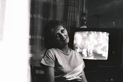 A young woman sits facing the camera, partially obscured by a bright window. She wears a graphic tee, seated near a vintage t...