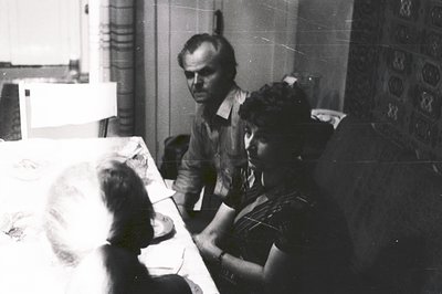 A man in a plaid shirt observes a woman seated at a table covered with a patterned cloth. A partial view of a young child is ...