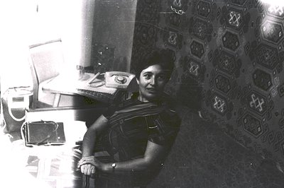 A seated woman in a patterned, sleeveless garment smiles directly at the camera. She is positioned within a room with pattern...