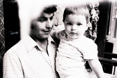 Man holds toddler, both smiling slightly. Interior setting features dark wood paneling & foliage. Casual attire suggests a do...