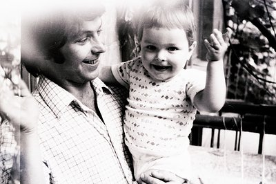 A smiling man holds a baby aloft, both gazing towards the camera. The man wears a plaid shirt; the baby, a patterned onesie. ...