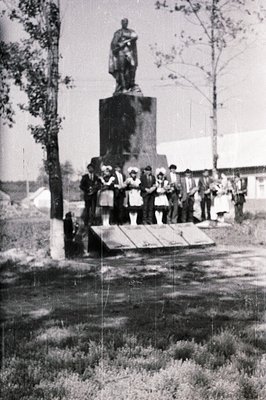 Monument stands in a village square with a group of formally dressed children and adults gathered at its base. The statue dep...