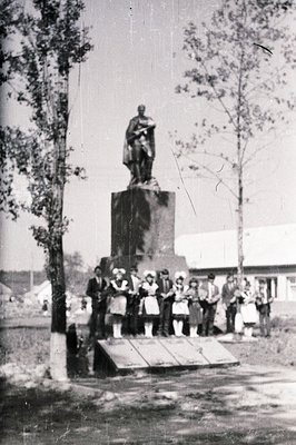 Monumental bronze statue atop a stone pedestal, viewed from a low angle. A group of formally-dressed children and adults stan...