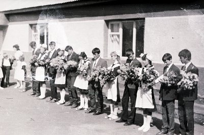 Group portrait of students, likely a graduating class. Boys wear dark suits & ties, girls wear white dresses with aprons. Eac...