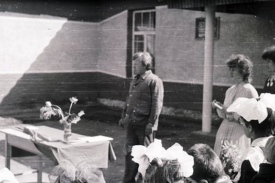 Black and white photo depicting a group of children, likely in a schoolyard setting. A man stands near a table draped with fa...