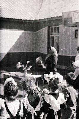 Group of children, likely school attendees, wear floral headwear and aprons in an outdoor courtyard setting. An adult figure ...