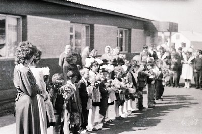 A line of schoolchildren, many holding flowers, stands before a low-slung building with large windows. A woman in a long dres...