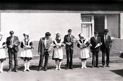 A group of six young people pose outside a simple, modernist building. Four wear traditional folk costumes with white aprons ...