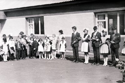 Group portrait outside a blocky, modernist building. A large group of children, some in uniform, stand alongside three adults...