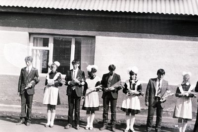 Six people pose in front of a building with a visible roof overhang and a single window. Four individuals wear vintage-style,...