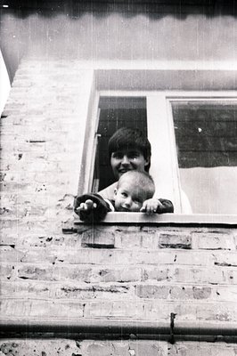 A young woman and baby peer out from a window with a brick facade. The woman has short hair and a cheerful expression; the ba...