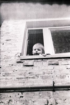 A young child peers from a window, framed by a brick wall and wrought-iron detail. Appears to be a candid moment, possibly a ...