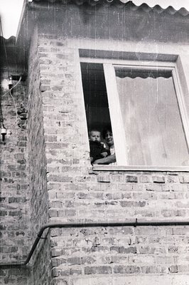 A young mother holding an infant sits posed in a window of a brick building. The exterior wall is visible, revealing aged bri...
