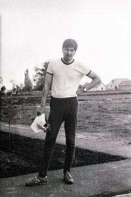 Young man stands outdoors, posed with one hand on hip, holding a folded cloth. He wears a white athletic tee, black trousers,...