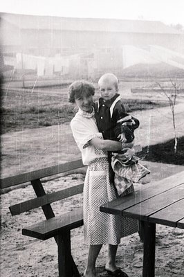 A woman in a patterned dress cradles a young boy on her hip, posed near a picnic table. Simple architecture and what appears ...