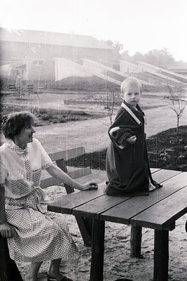 A young boy stands atop a wooden picnic table during a rain shower, bundled in a dark coat. A woman, seated beside him, obser...
